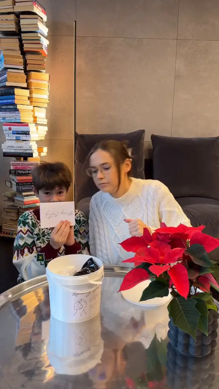 Mama su kūdikiu namų kambaryje, knygų lentynų fone.